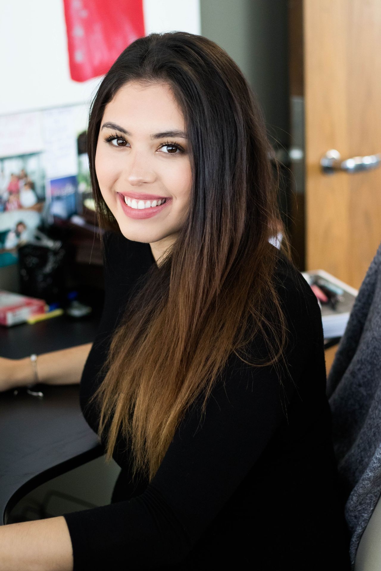 Smiling person with long hair sitting at a desk in an office setting.